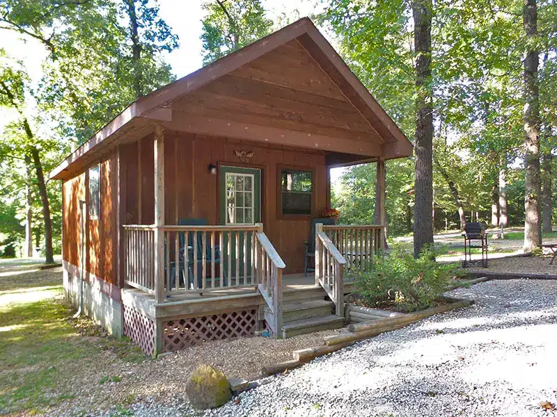 Small wooden cabin with covered front porch, railing, chairs, and steps, set among tall trees beside a gravel drive.