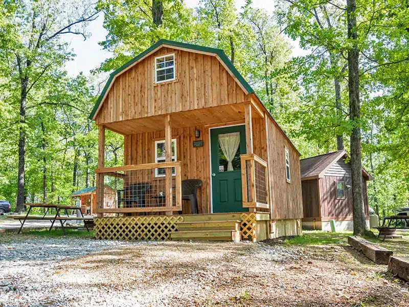 Two-story cedar camping cabin with green metal roof, covered porch and green door, with picnic table on gravel among