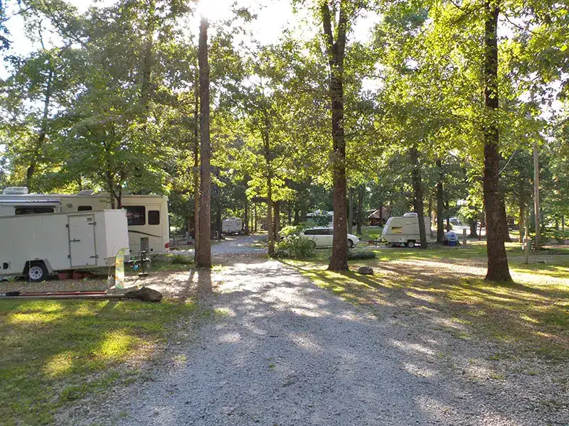 Gravel campground road winding between shaded RV sites with parked travel trailers, a white car, and tall leafy trees.
