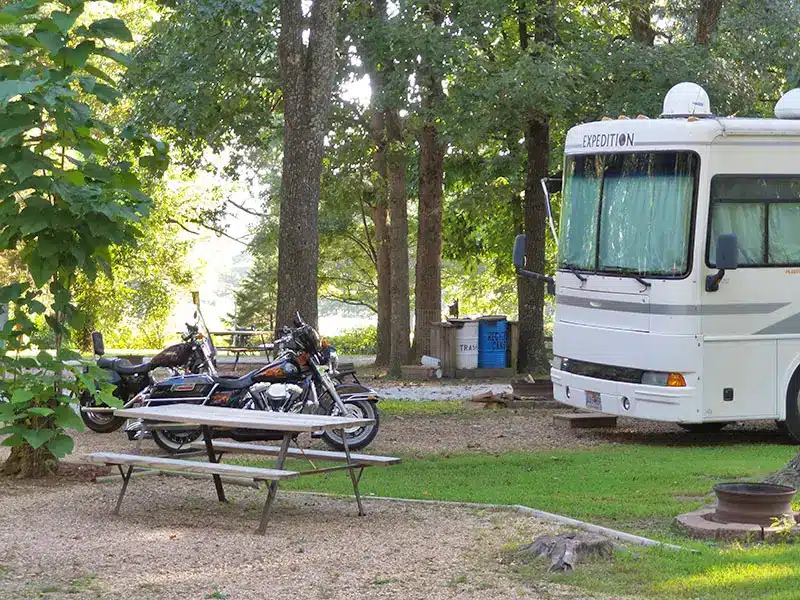 Class A Expedition motorhome parked at a shaded gravel site with two motorcycles, a wooden picnic table, and fire ring.