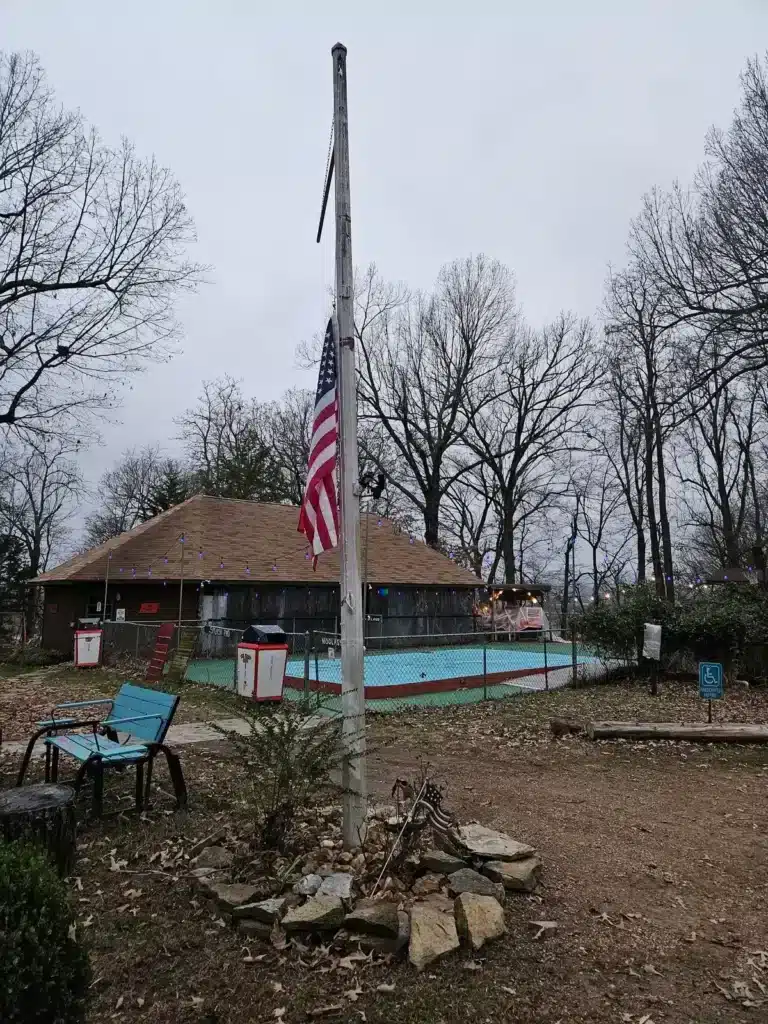American flag on a tall pole beside a fenced empty turquoise swimming pool and shingled clubhouse among bare winter