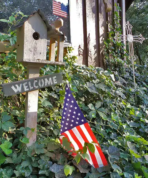 Rustic wooden birdhouse atop a post with a 'Welcome' sign, American flag banner, and decorative cross amid ivy.